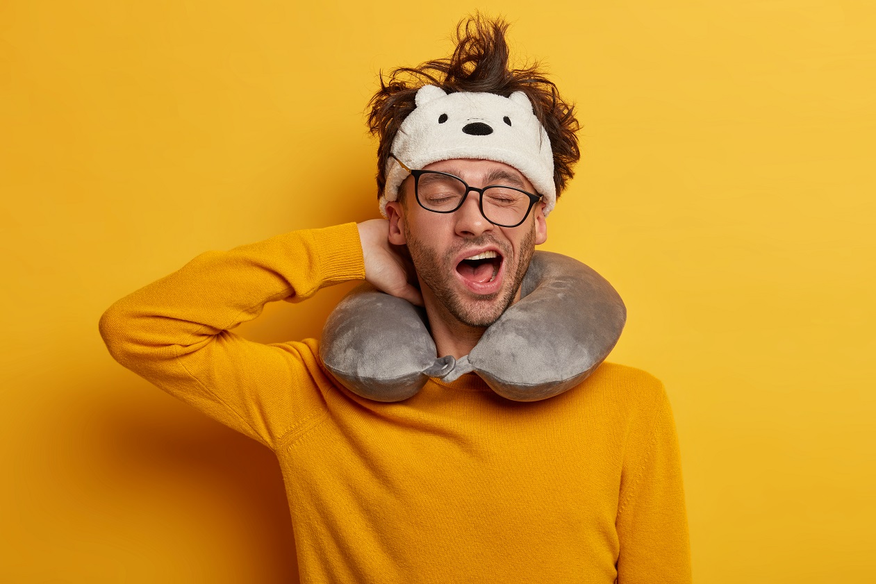 sleepy male traveler yawns as wants to sleep, travels in transport with neck pillow, wears transparent glasses, headband, yellow jumper, poses indoor. people, tiredness and travelling concept