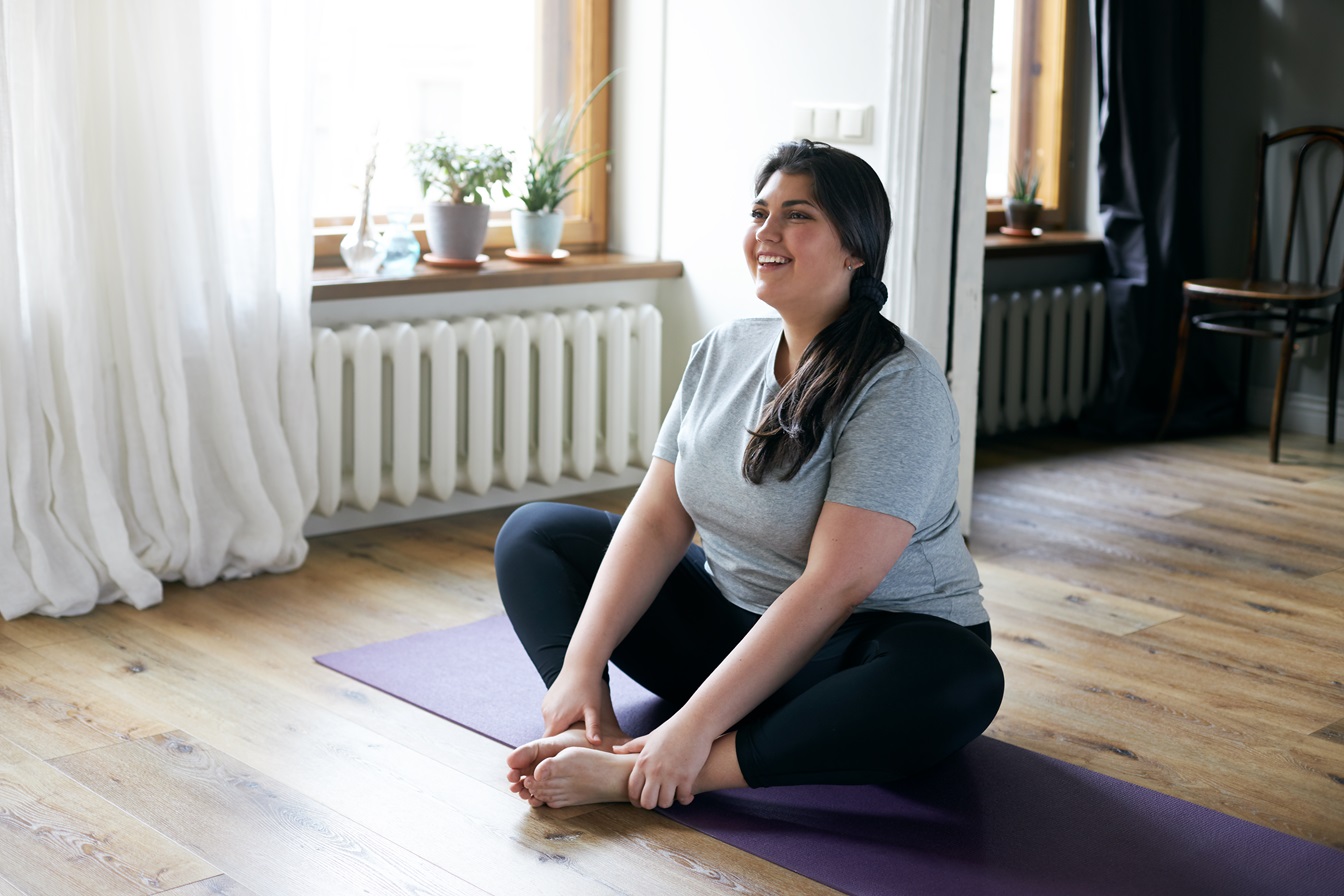 cheerful attractive young overweight woman in activewear choosing healthy lifestyle, sitting on mat with hands on bare feet, doing butterfly yoga exercise, stretching thighs. body shape and activity