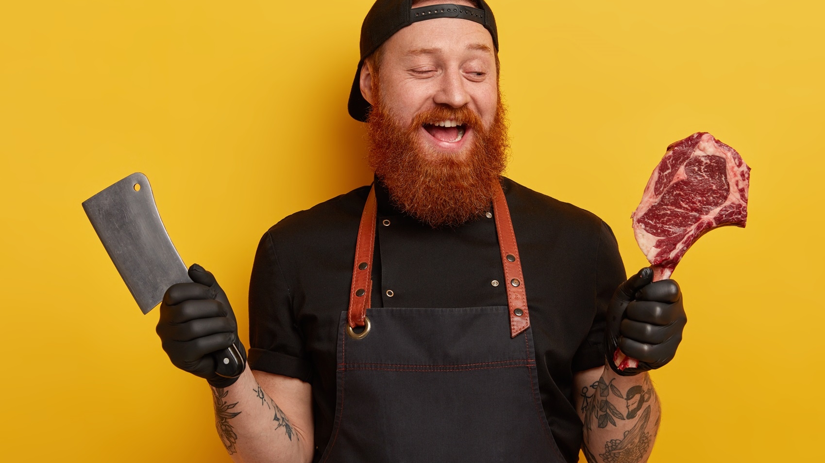 happy foxy bearded man sells meat with bone in butcher shop, cuts with cleaver on pieces, wears black cap, gloves and apron, isolated over yellow wall. people, production, nutrition concept.