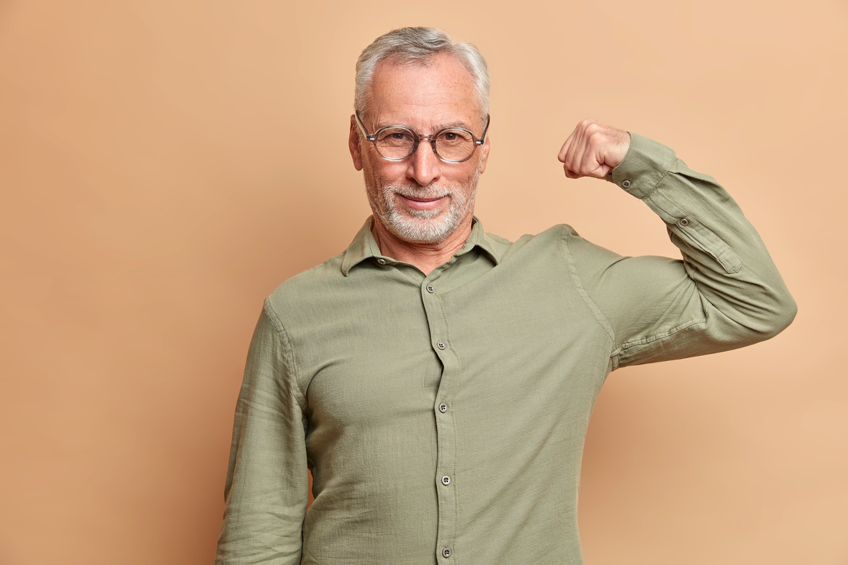 self confident serious man raises arm shows muscles being confident in his strength wears formal shirt poses against brown background. healthy handsome european pensioner demonstrates biceps