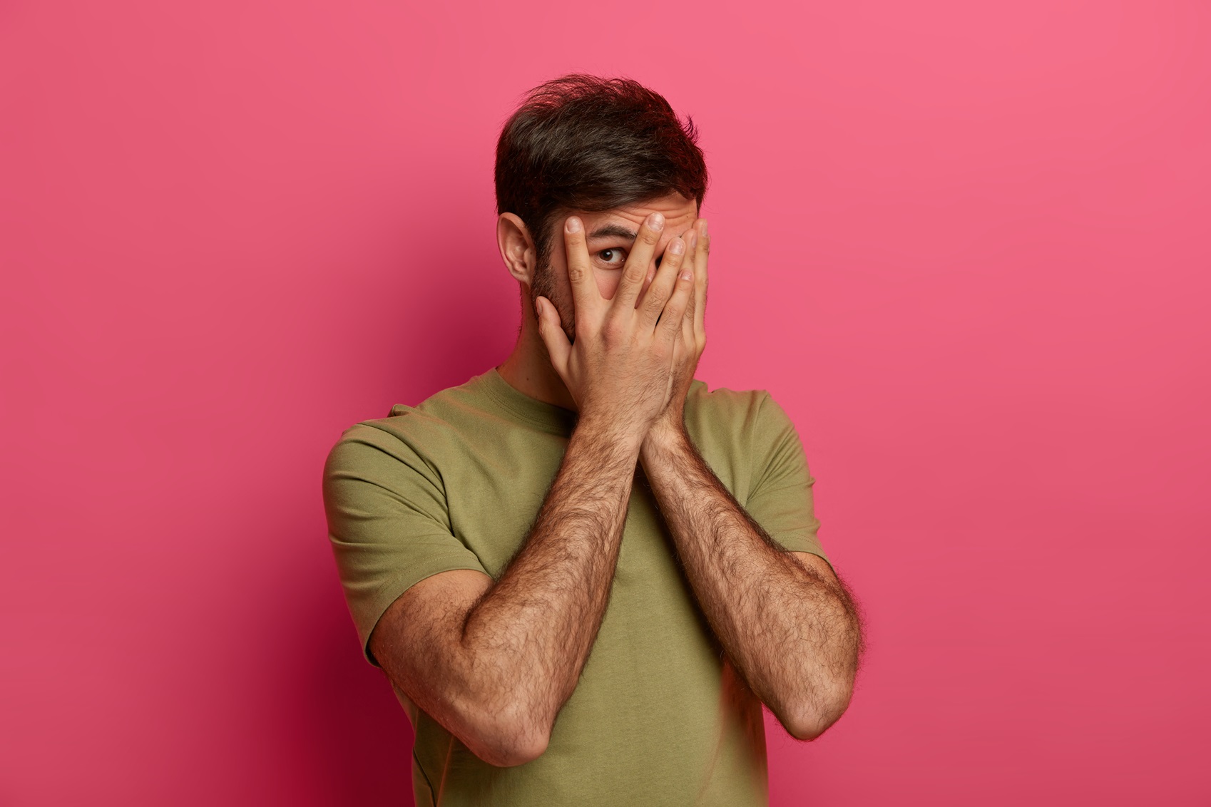 surprised european man covers face with hands, looks through fingers, hides himself, has mysterious expression, wears casual t shirt, poses over rosy studio background. dont look at me, please